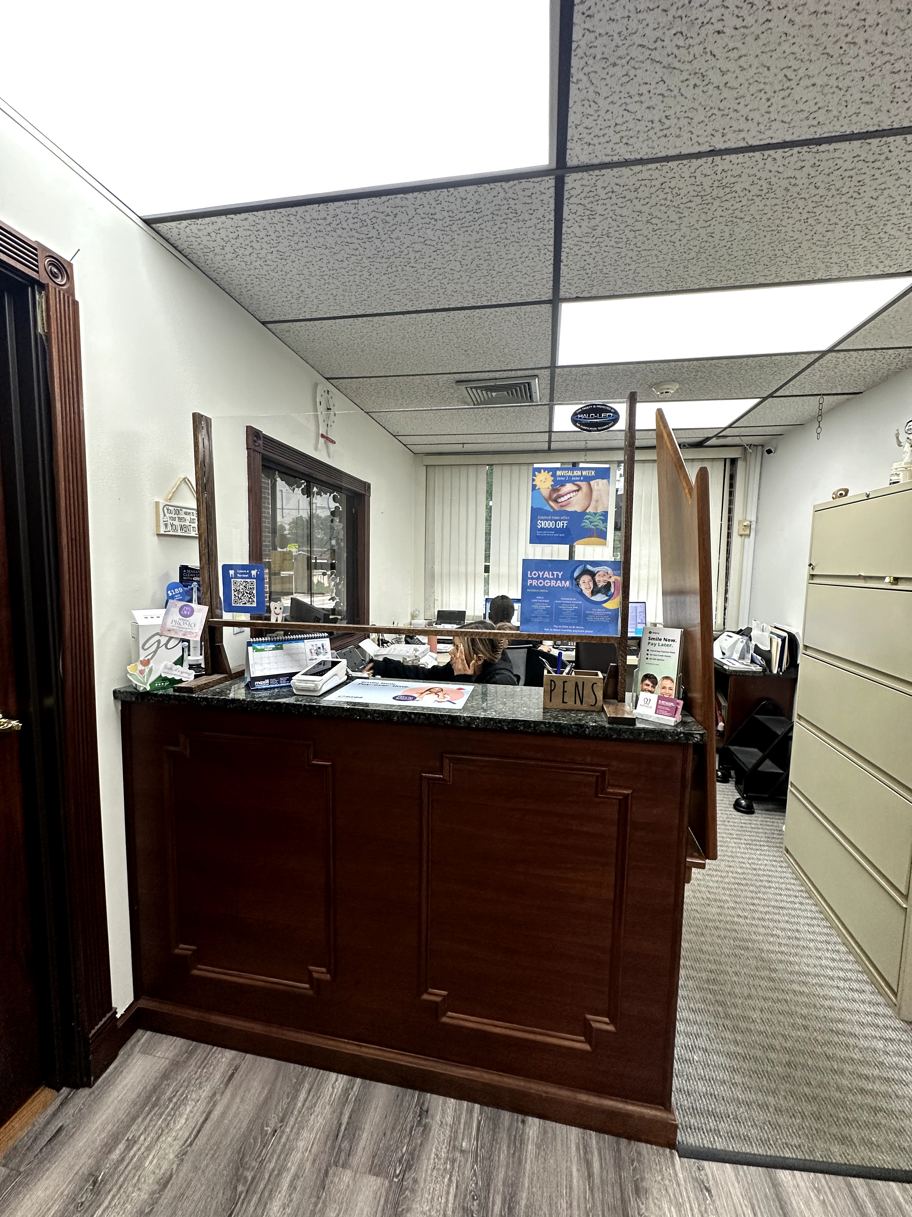 Reception area with dark wood desk, granite-look counter, drop ceiling, and mixed flooring in Rochelle Park, NJ showing dated commercial finishes needing renovation.