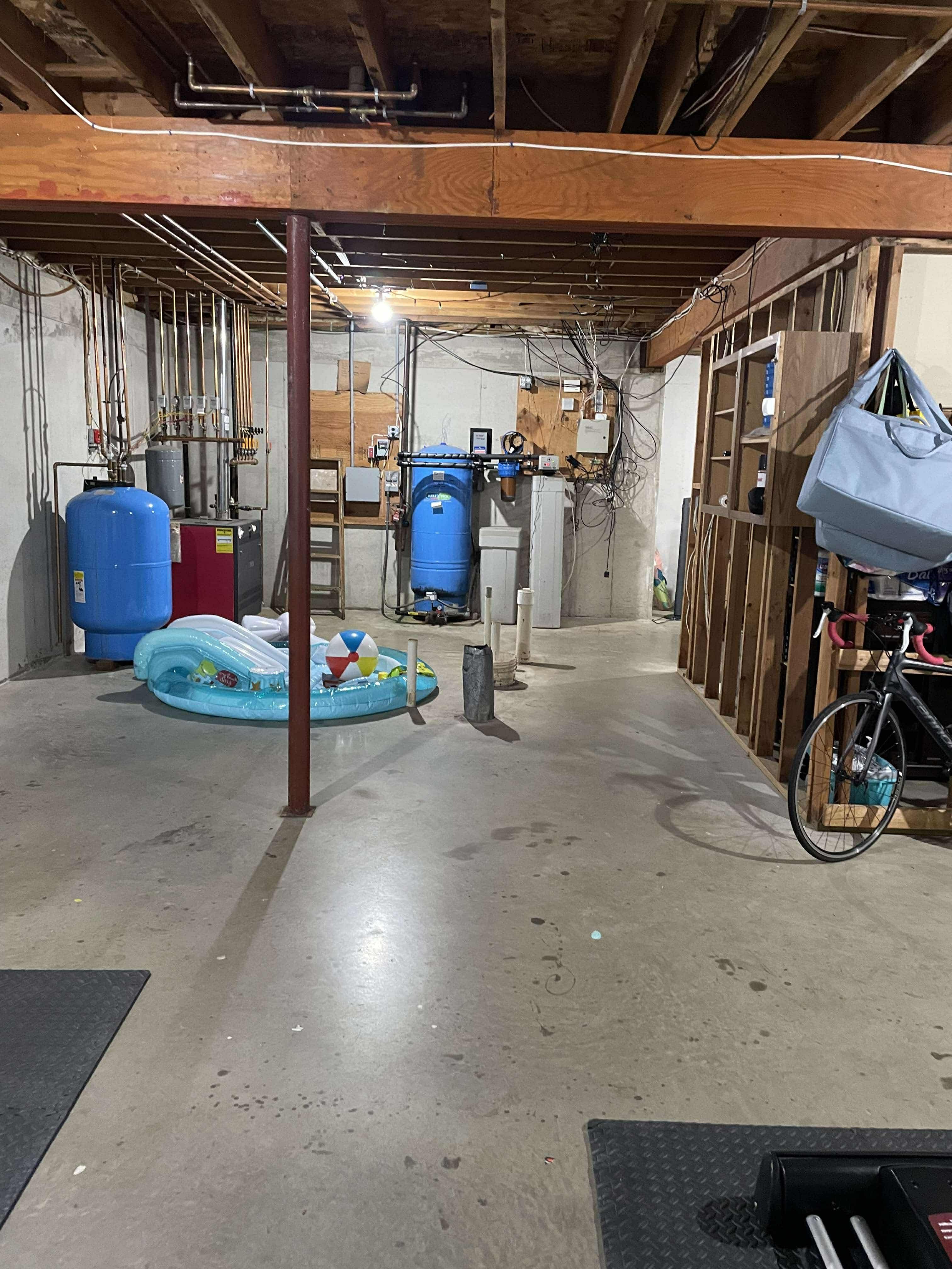 Unfinished basement with concrete floor, exposed wood beams, utility pipes, and visible wiring in Kinnelon, NJ before renovation.