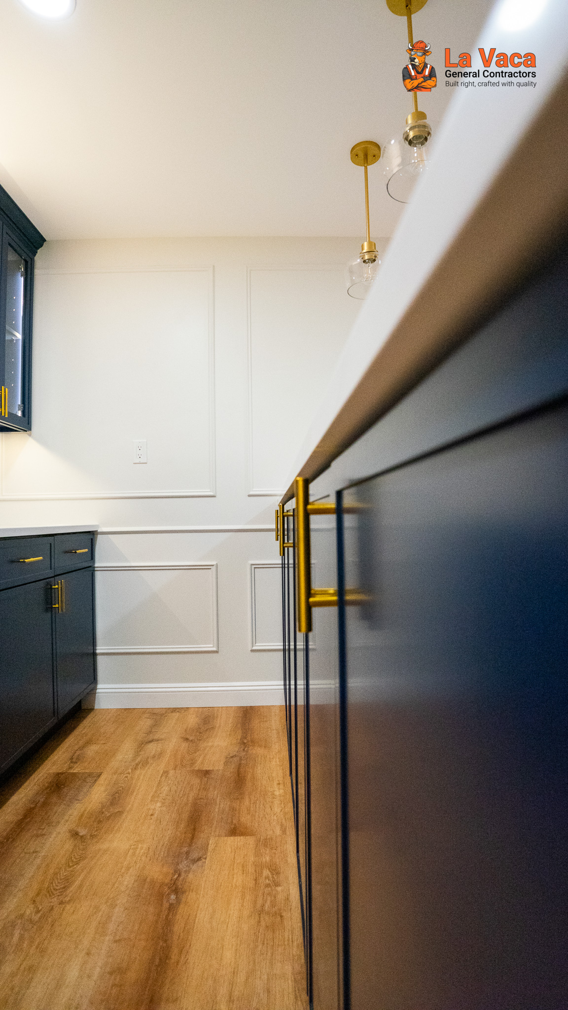 Basement bar featuring navy cabinets, gold hardware, wood-look flooring, and white paneled walls after Basement Finishing in Kinnelon, NJ.