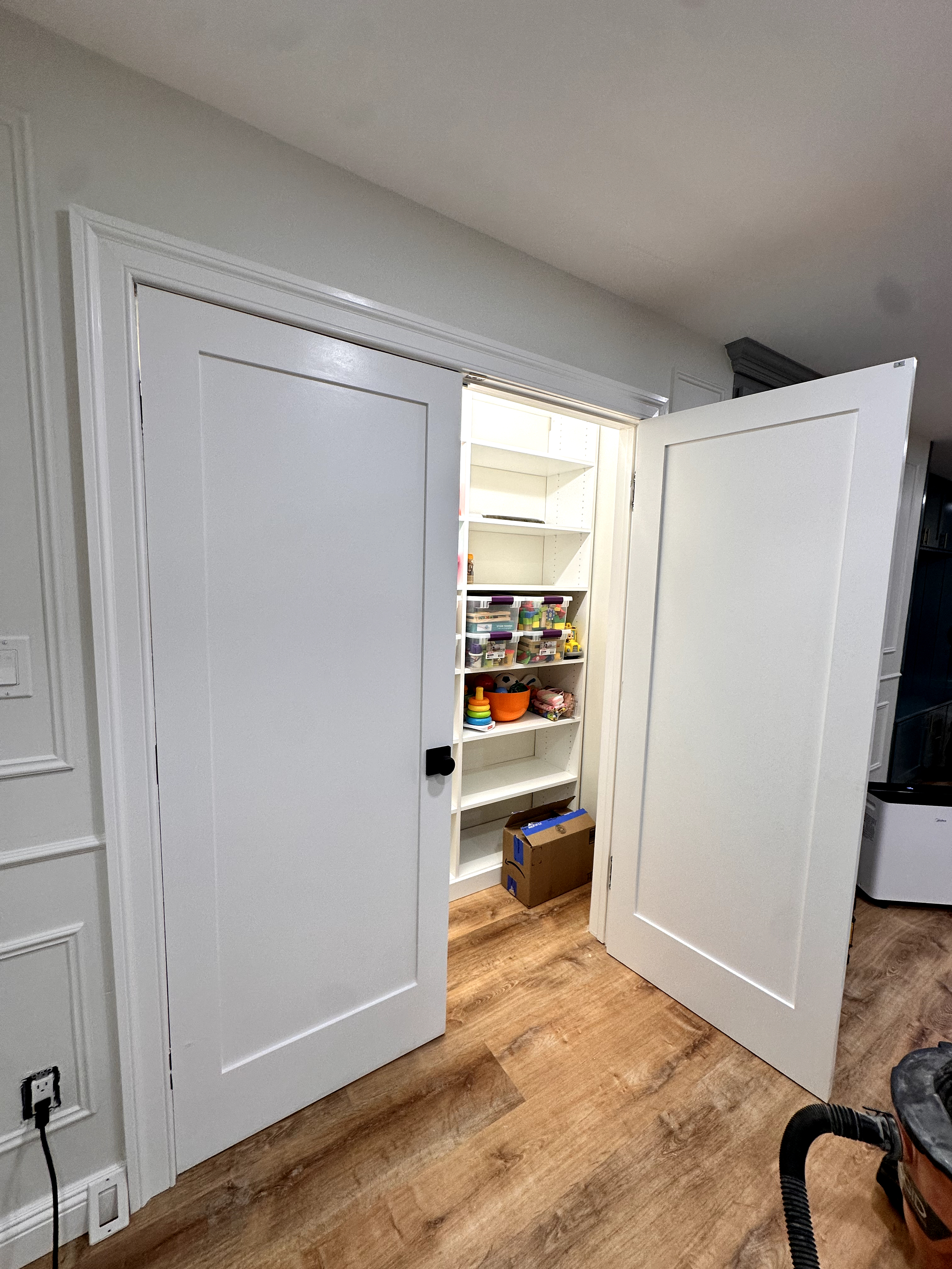 Basement pantry with white flat panel double doors, wood-look laminate flooring, and built-in white shelving after Basement Finishing in Kinnelon, NJ.